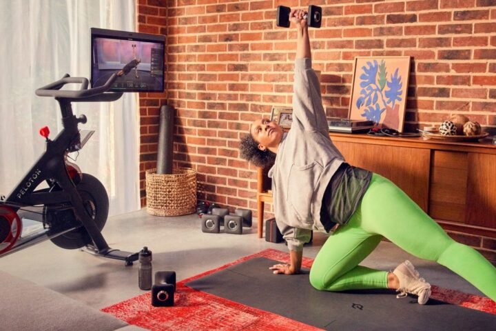 Woman performing dumbbell exercise on yoga mat. A Peloton Bike+ is next to her with the in-built screen turned her way and showing a fitness class
