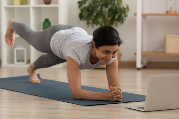 Woman performs plank with leg lift on an exercise mat in her living room, a laptop is open in front of her
